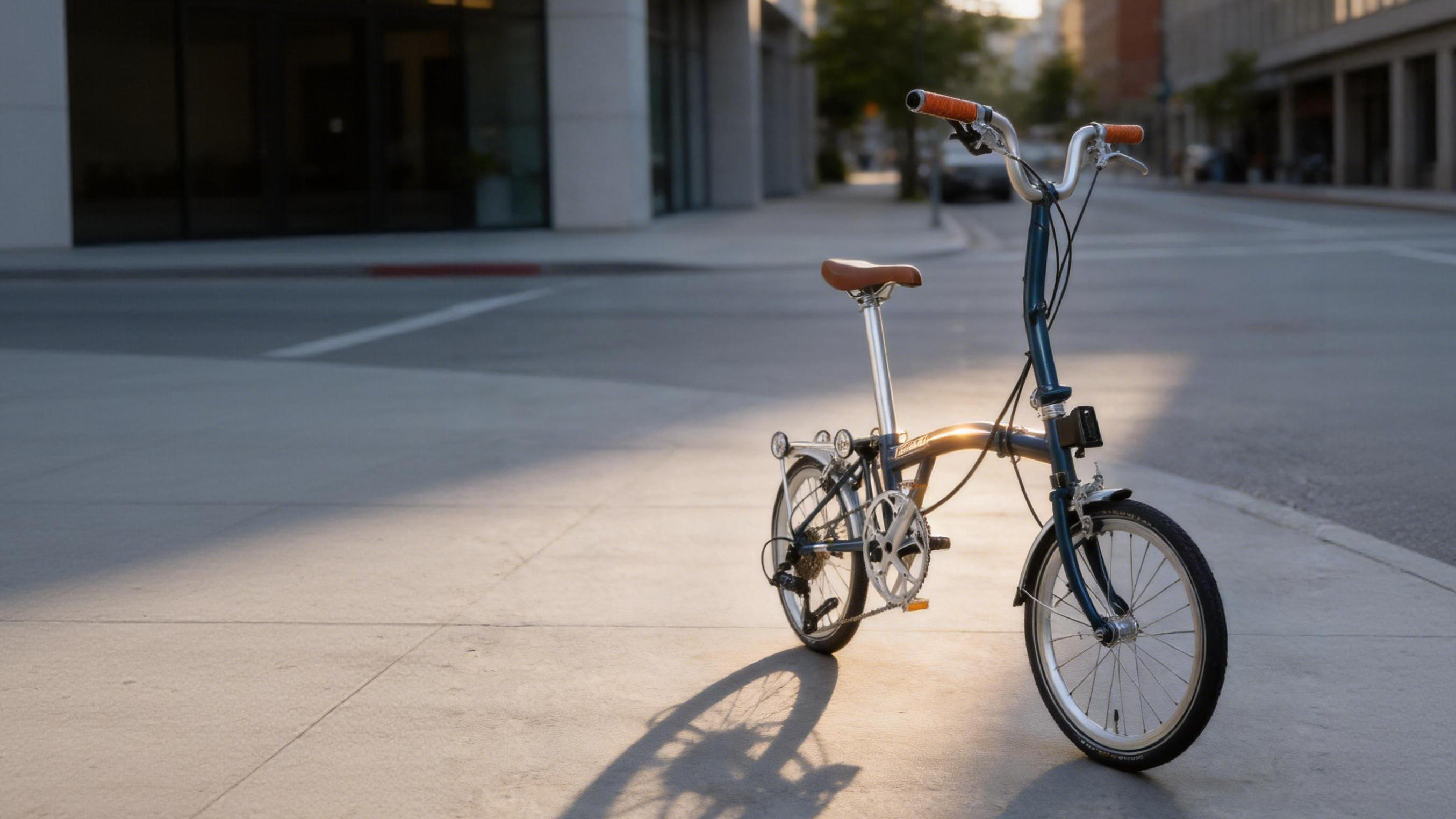 A folding bicycle with a blue frame, brown saddle and handlebar grips, and silver wheels, parked on a city sidewalk between modern buildings during late afternoon sunlight.